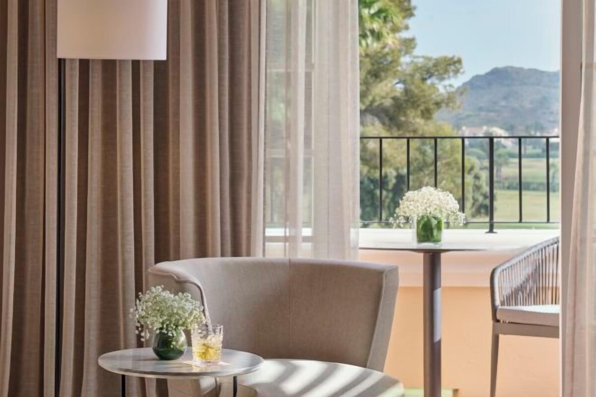 Ein Balkon mit Tisch und Stuhl, eine Vase mit Blumen und Blick auf Grünflächen und Hügel.