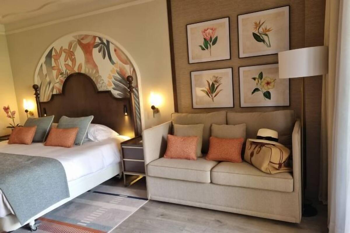 Una habitación de hotel con cama, sofá, mesita de noche, lámpara y decoración floral en la pared.