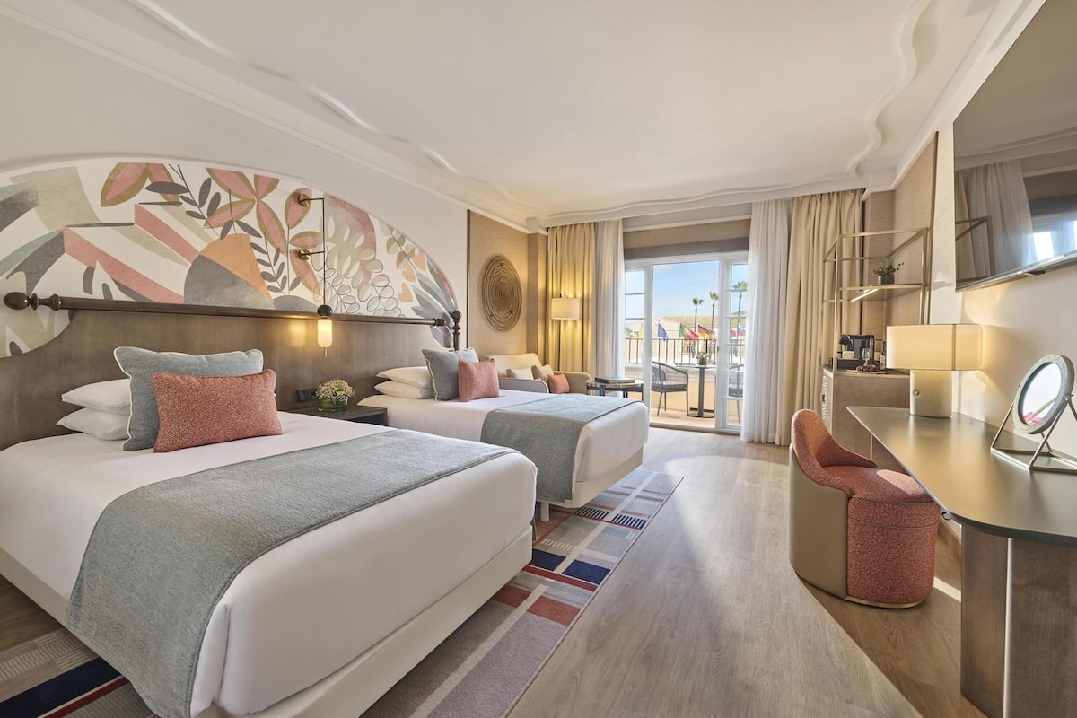 Ein Hotelzimmer mit zwei Betten, einem Schreibtisch, einem Stuhl, einem großen Fenster und Blick auf den Strand.