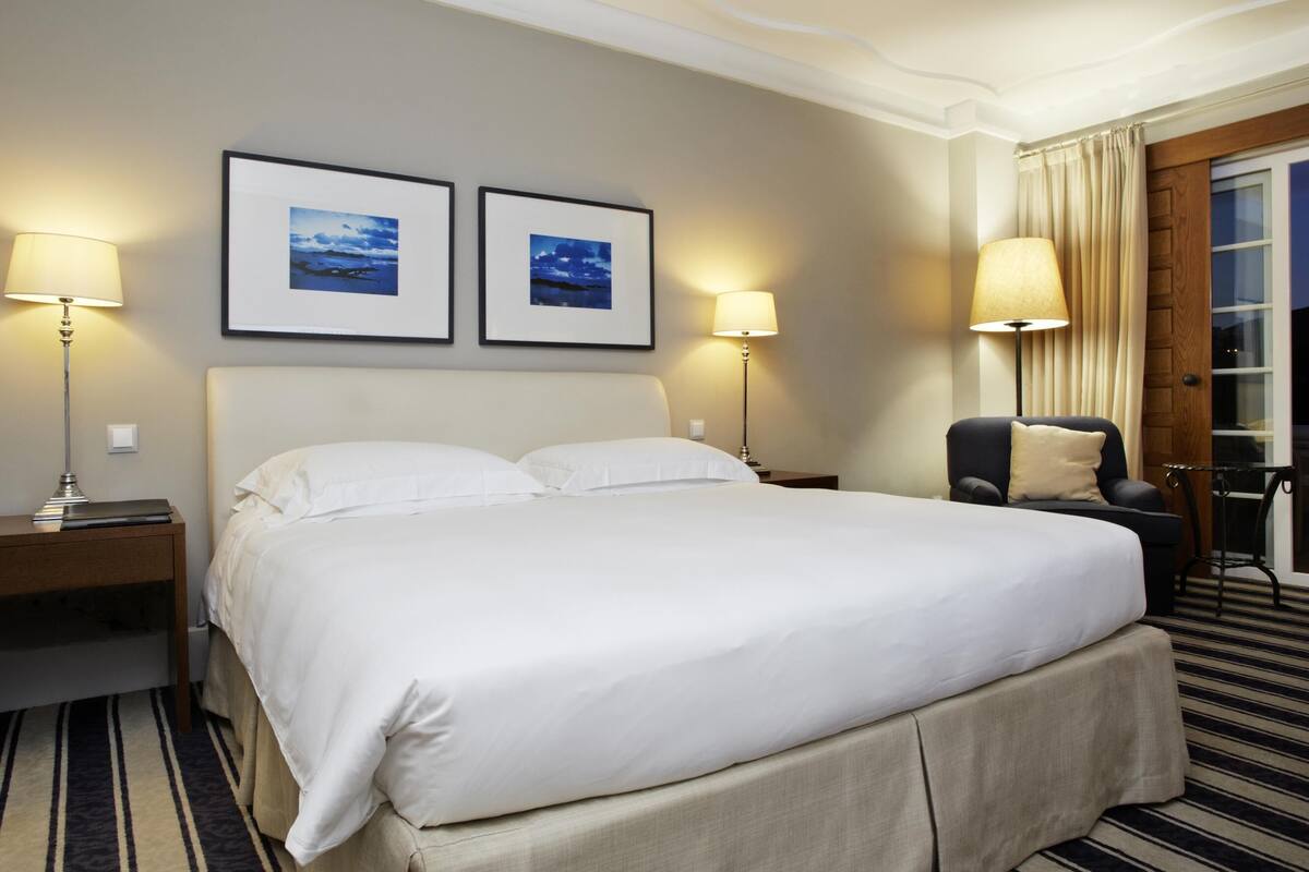 Una habitación de hotel con una cama grande, dos lámparas de noche, una silla y dos cuadros en la pared.