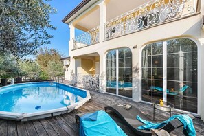 Une piscine extérieure dotée d’une terrasse en bois, de chaises longues et d’une grande maison avec des balcons décoratifs et de grandes fenêtres.