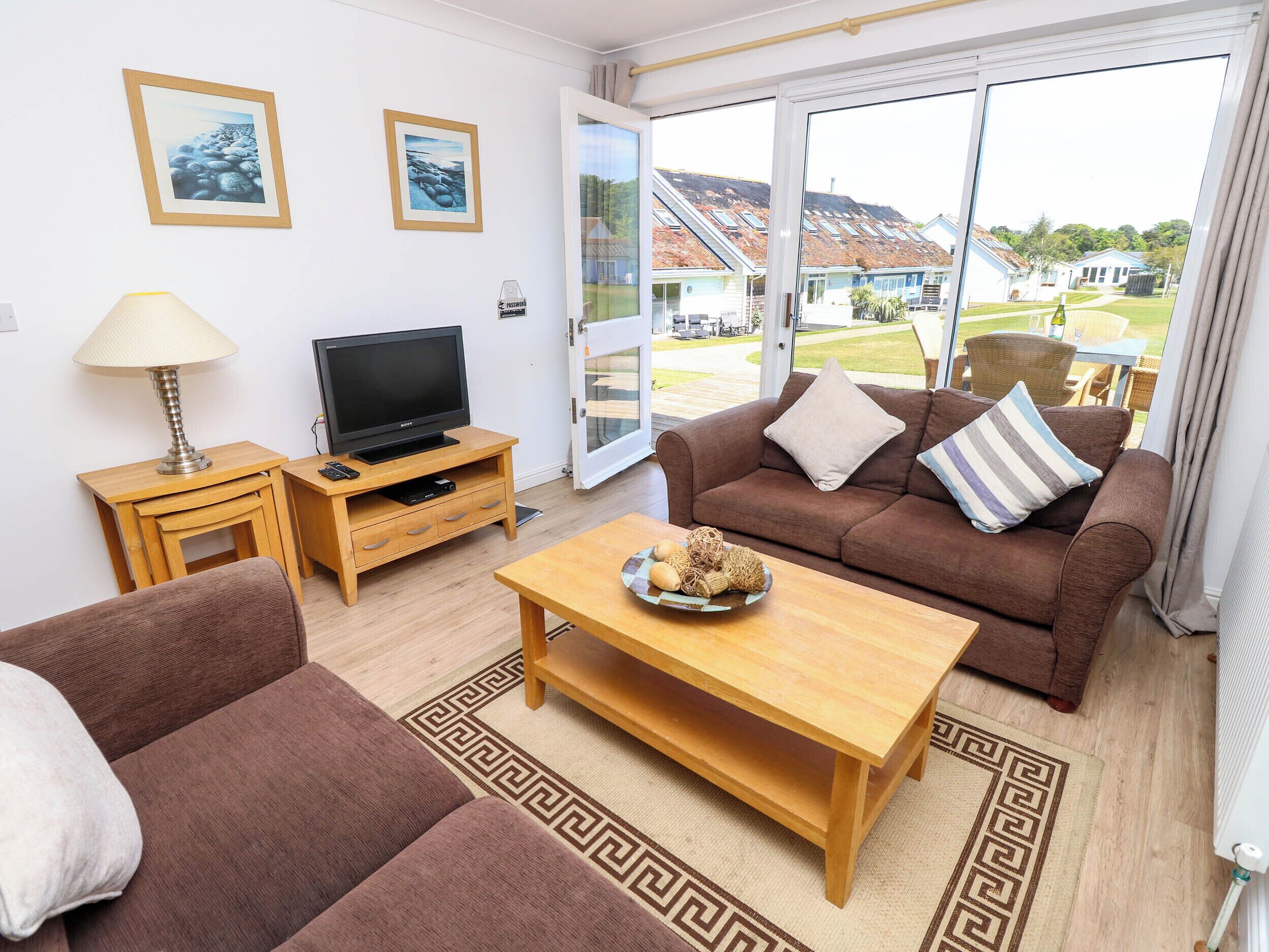A furnished living room with a sofa, a coffee table, a TV on a wooden stand, and a view of a residential area through a glass door.