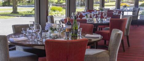 Een restaurant met ronde tafels, rode beklede stoelen en uitzicht op een tuin door grote ramen.