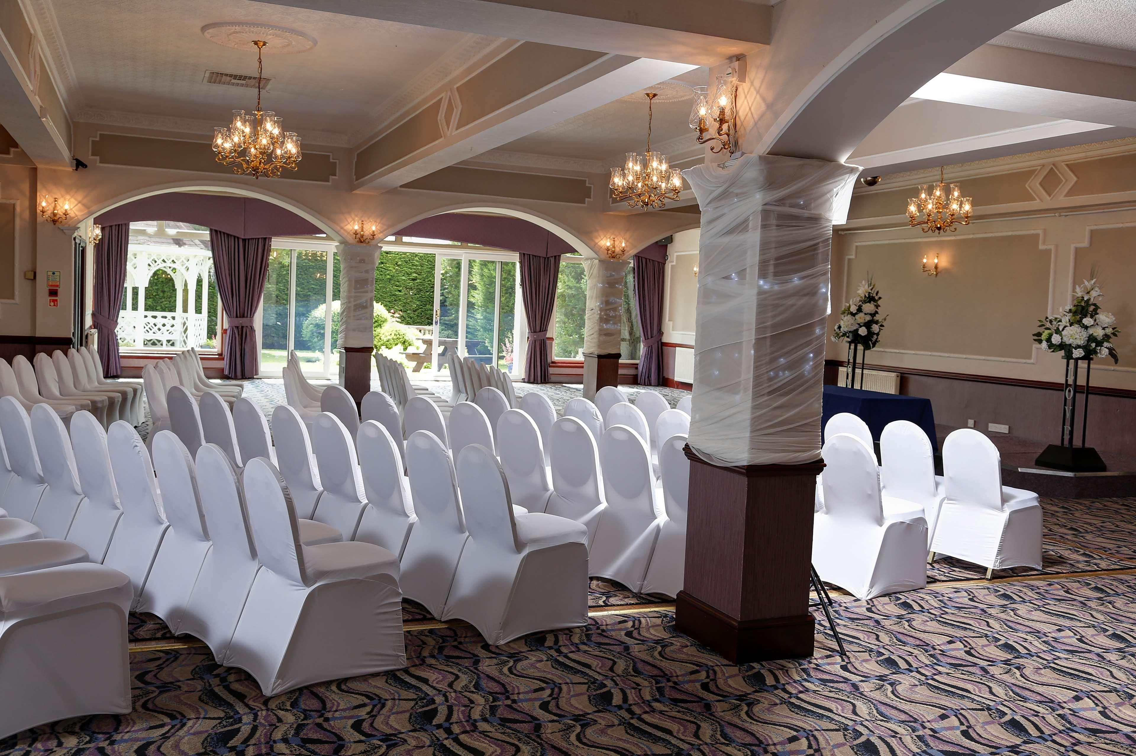 A conference room with rows of white chairs, chandeliers, and a large screen.