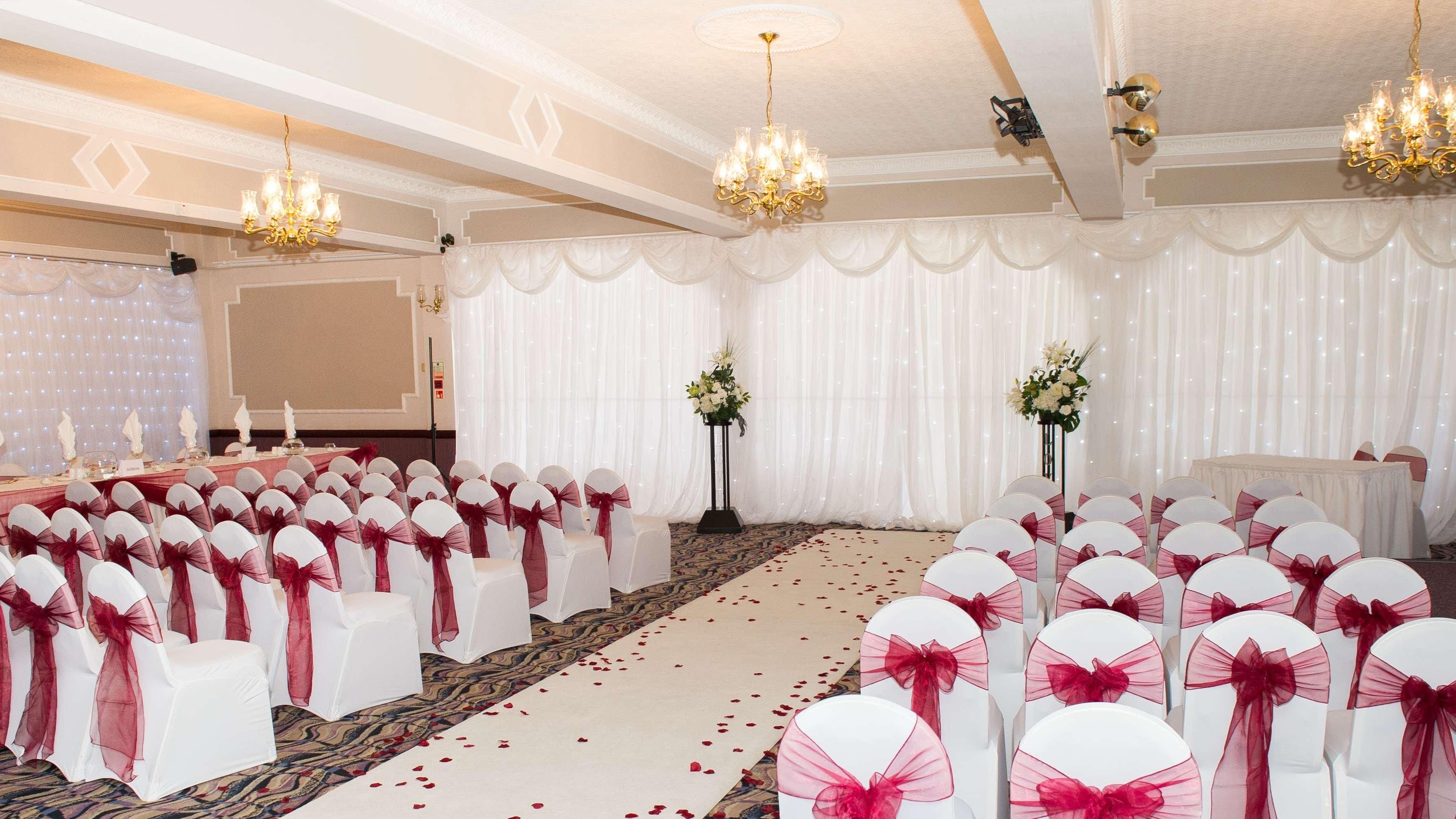 A banquet hall with rows of white chairs with red sashes, chandeliers, and floral centerpieces.