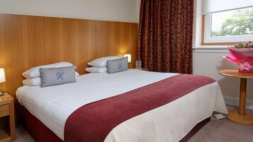 A hotel room with a large bed, two pillows with a monogram, a bedside table with a lamp, a window with curtains, and a small table with a bouquet.