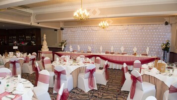 Un salón de banquetes con mesas redondas, sillas blancas con lazos rojos, un escenario con cortinas y un candelabro.