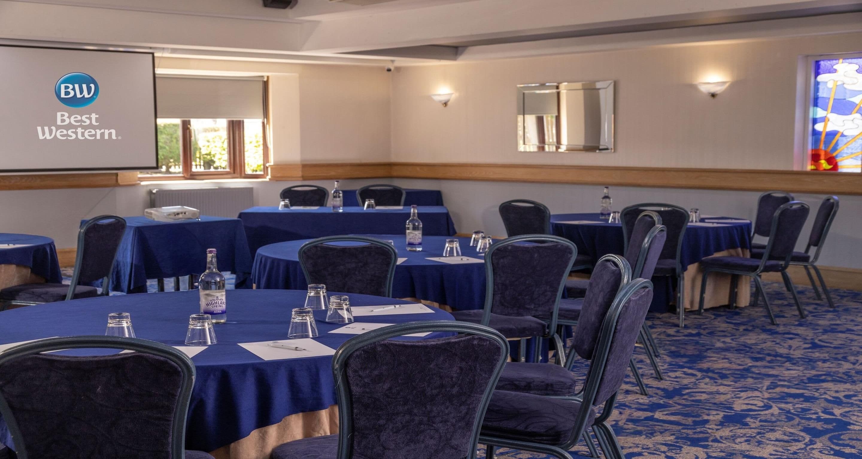 Un salón de conferencias con mesas redondas, sillas y una pantalla de proyector mostrando 'BW Best Western'.