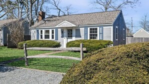 A single-story house with a gabled roof, a front porch, and a small front yard with a wooden fence and trimmed bushes.