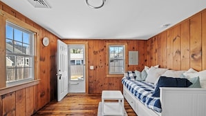 A cozy room with wooden walls, a bed with a checkered bedspread, a white bench, and a window with a view of a house.