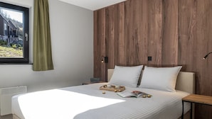A bedroom with a large bed, wooden panel wall, a window with a curtain, bedside table with a lamp, and a tray with cups and a book.