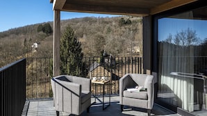 A balcony with two grey armchairs and a small table, overlooking a forested hillside.
