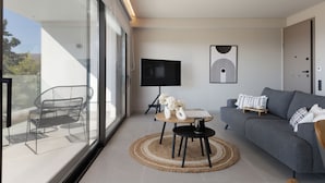 Una sala de estar moderna con un sofá gris, un televisor sobre un mueble bajo, una mesa redonda con un jarrón y una taza, y una silla junto a un amplio ventanal.