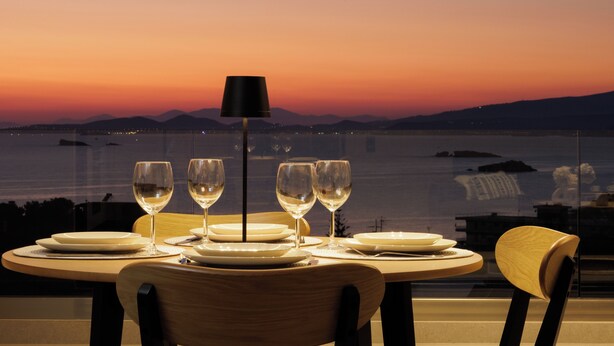 Mesa para cuatro personas con copas de vino y platos, con vista a una masa de agua al atardecer.
