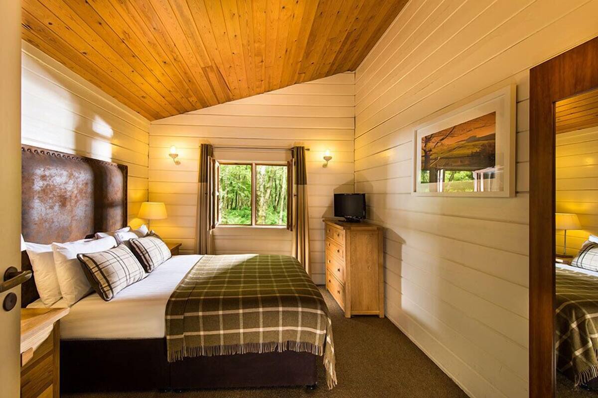 Ein gemütliches Schlafzimmer mit Holzbalkendecke, einem Bett mit karierter Decke, einem Nachttisch mit Lampe und einem Fenster mit Vorhängen.