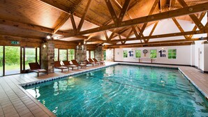 An indoor swimming pool with wooden beams, lounge chairs, and a stone wall.