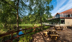 Een terras aan de rivier met houten tafels en banken, een houten hek en een gebouw met een rood pannendak.
