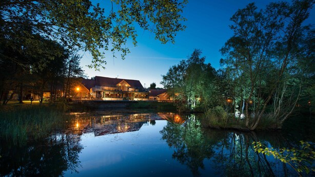 A lakeside hotel with a reflection in the water, surrounded by trees and a well-lit building.