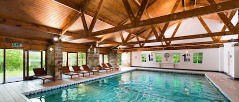 An indoor swimming pool with wooden beams, lounge chairs, and a stone wall.