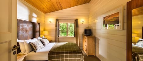 Une chambre confortable avec un plafond en bois, un lit recouvert d’une couverture à carreaux, une table de chevet avec une lampe et une fenêtre avec des rideaux.