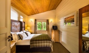 Een gezellige slaapkamer met een houten plafond, een bed met een geruit plaid, een nachtkastje met een lamp en een raam met gordijnen.