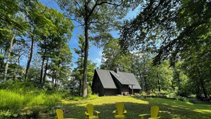 A cabin surrounded by trees with a fire pit and three yellow Adirondack chairs in the foreground. - Woodland A-Frame (Sharon)