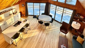 A cozy, wooden interior with a dining area, kitchen, and large windows offering a view of a snowy landscape. - Woodland A-Frame (Sharon)
