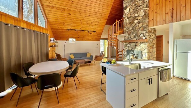 A modern kitchen with a stone accent wall, wooden ceiling, and a dining area with a round table and chairs. - Woodland A-Frame (Sharon)