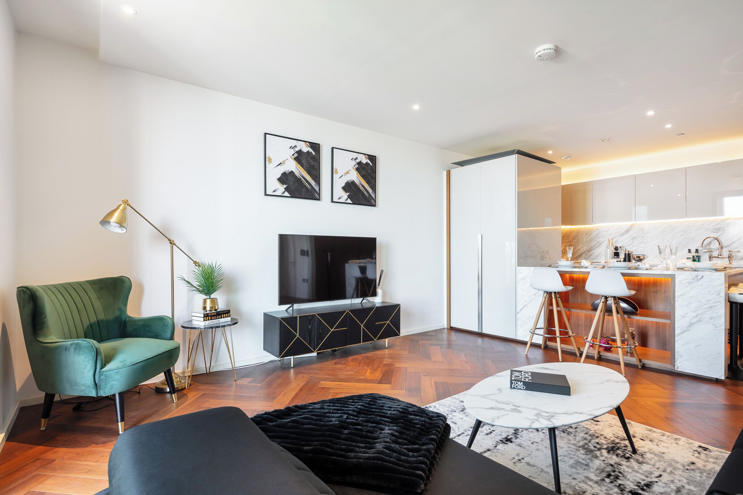 Un salon moderne avec un fauteuil vert, un pouf noir et un téléviseur posé sur un meuble noir. On y trouve également un coin cuisine avec un plan de travail en marbre et des tabourets.