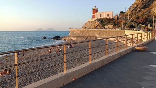A coastal promenade with a stone wall, a lighthouse, and a beach with people. - VACATION HOME SUN & BEACH - LEMONS APARTMENT (Capo d'Orlando)