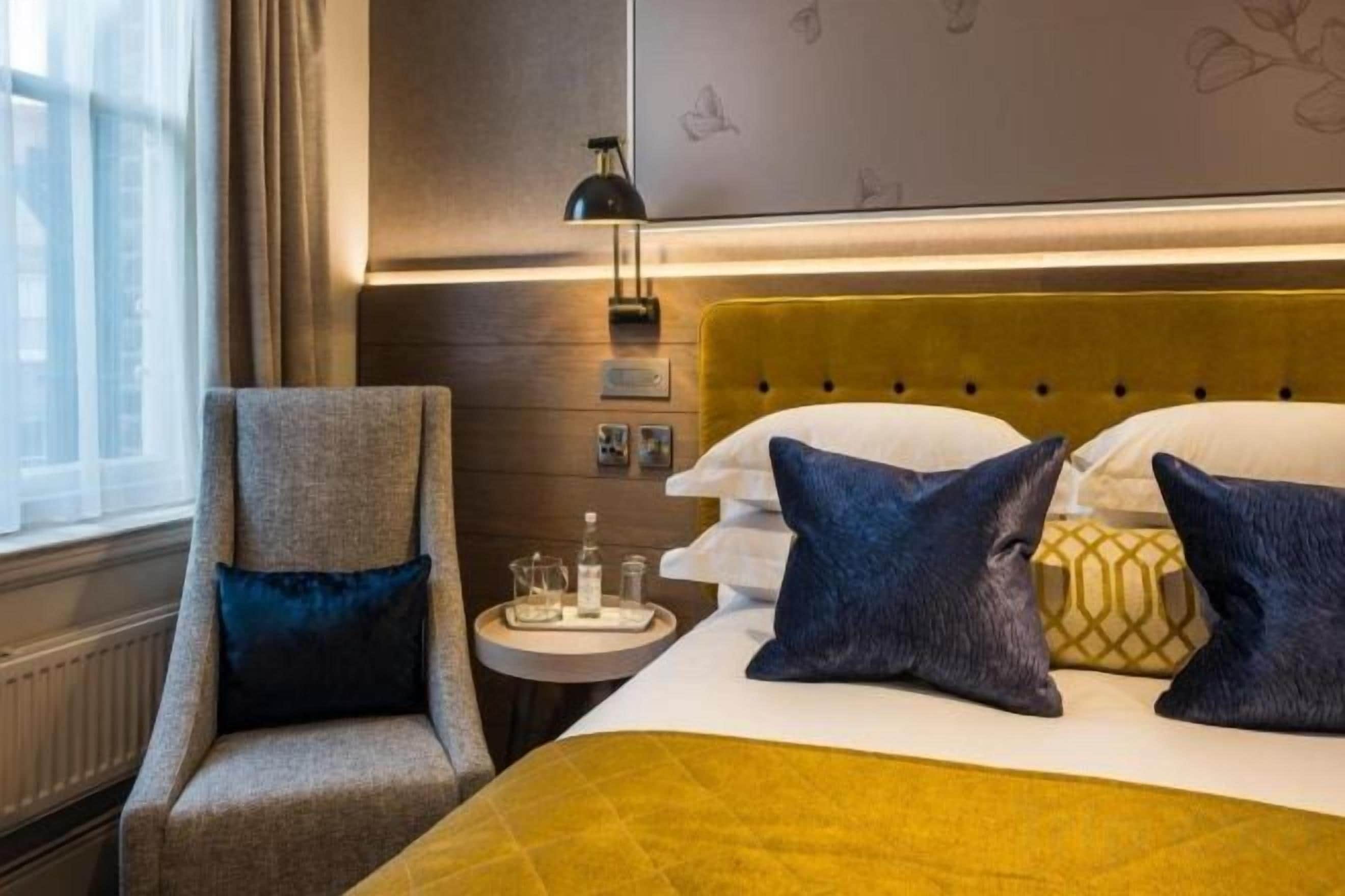 A hotel room with a bed, a chair, a bedside table, and a lamp.