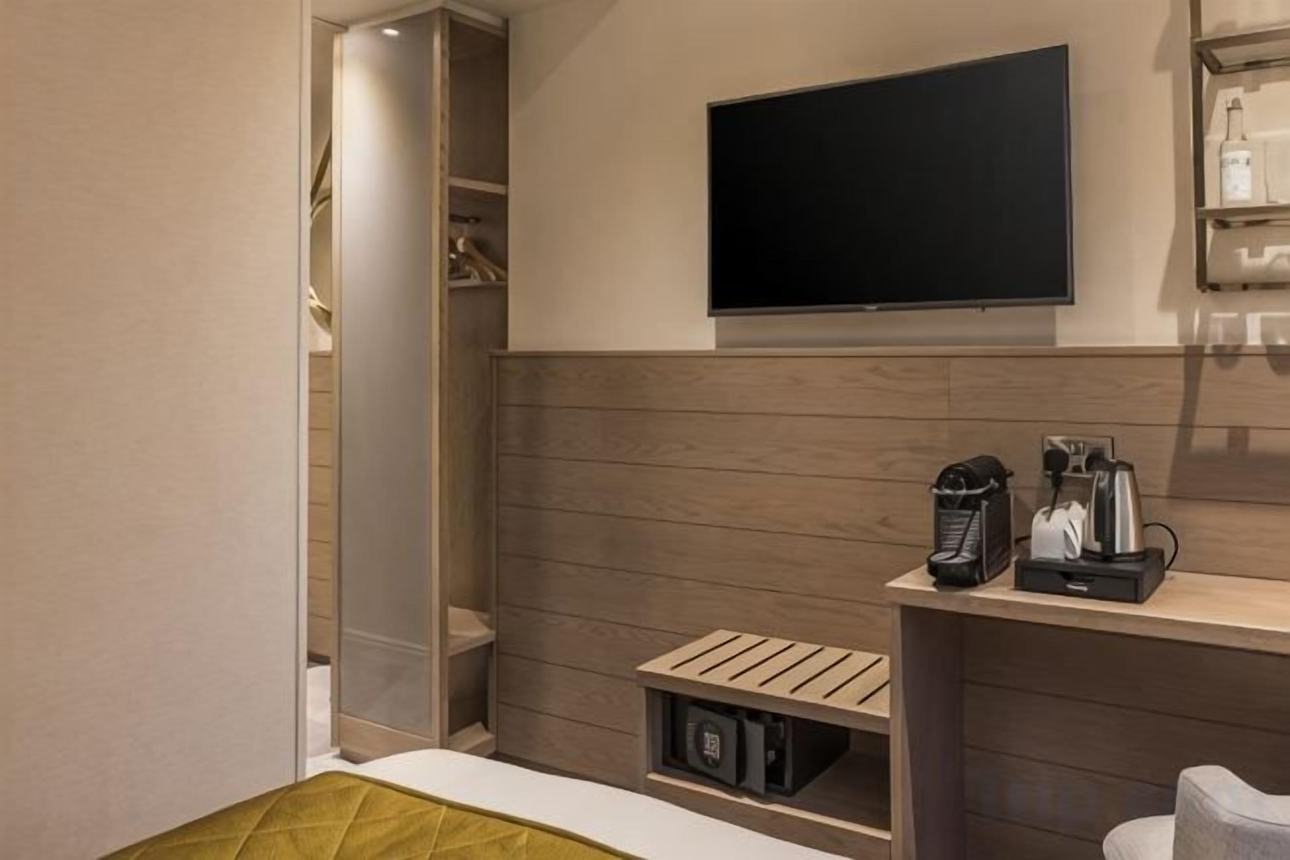 A hotel room with a bed, a TV mounted on the wall, a coffee machine, and a shelf with toiletries.
