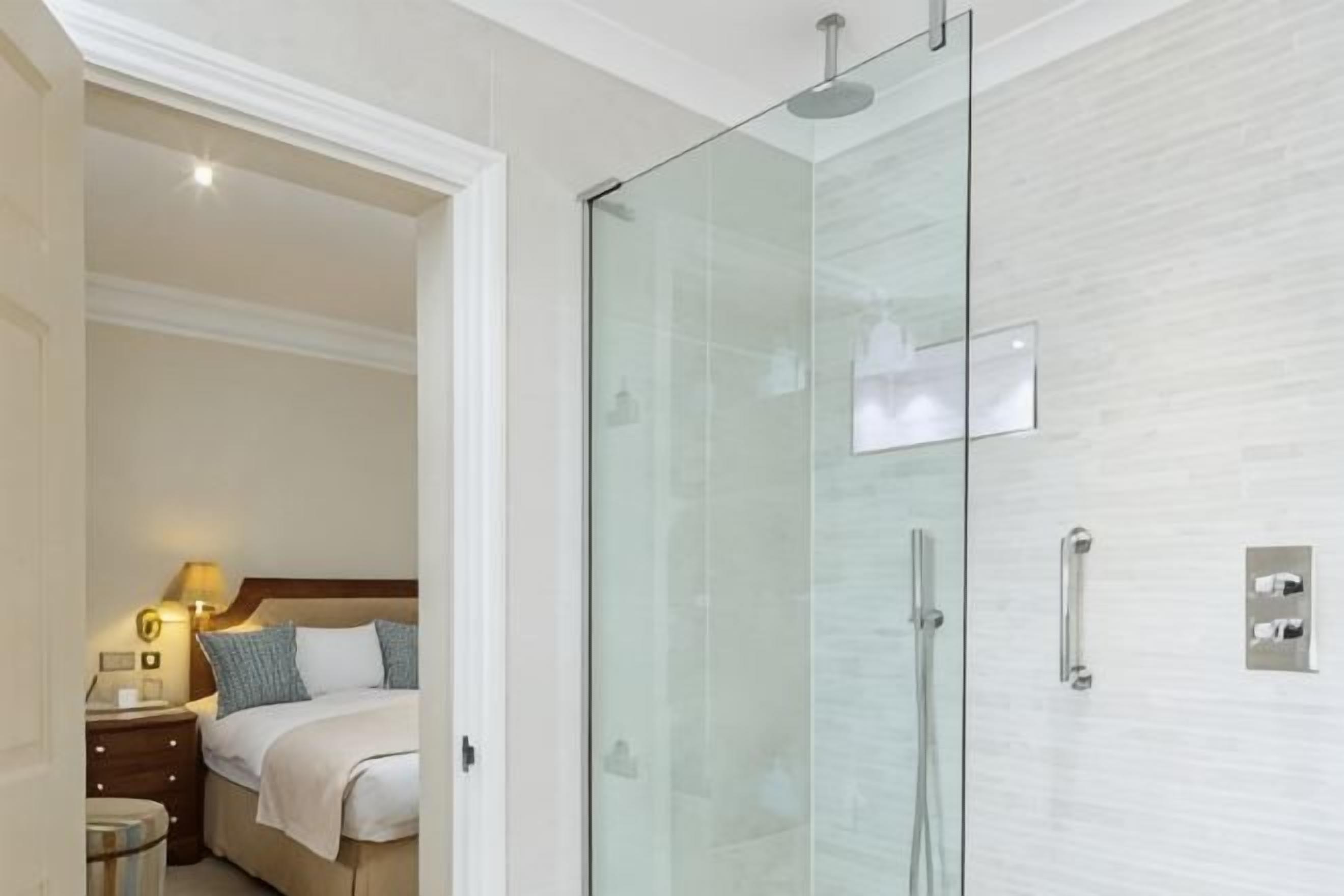 A hotel room with a glass-enclosed shower, a bed with pillows, a nightstand with a lamp, and a wooden headboard.