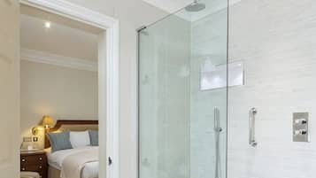 A hotel room with a glass-enclosed shower, a bed with pillows, a nightstand with a lamp, and a wooden headboard.