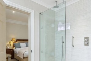 A hotel room with a glass-enclosed shower, a bed with pillows, a nightstand with a lamp, and a wooden headboard. - Sloane Place (London)