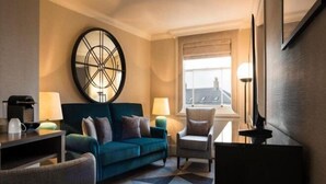 A hotel room with a sofa, armchairs, a coffee table, a desk, a lamp, a large wall clock, and a window with a view. - Sloane Place (London)