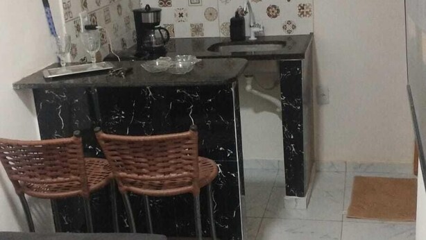 A compact kitchen with a black marble countertop, a coffee machine, and a shelf above the counter. There are three wicker chairs at the counter and a grey sofa in the living area. - Loft Pajuçara (Maceio)