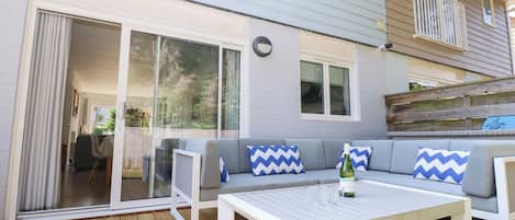 A covered patio with a white table, grey sofa with blue and white chevron cushions, and a bottle of wine.