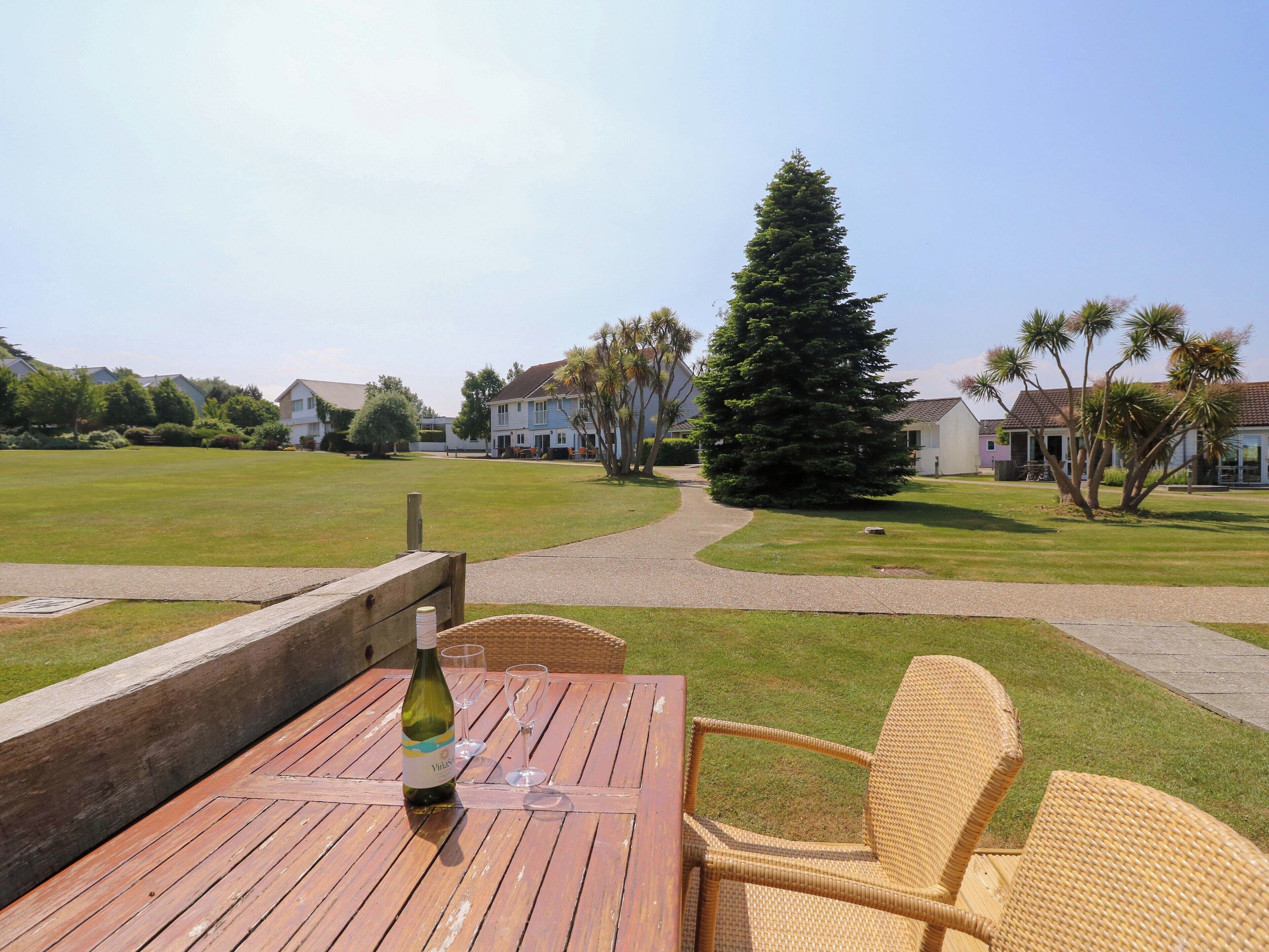A wooden table with a bottle and glasses, overlooking a well-maintained lawn and houses in the background.