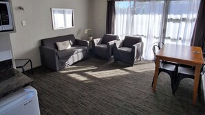 A modern living room with a sofa set, a dining table with chairs, a small kitchen area with a refrigerator, and large windows with curtains. - cherry grove motel (Havelock North)