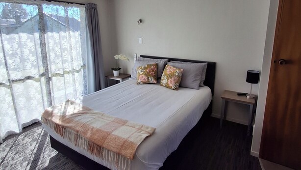 A hotel room with a large bed, a bedside table with a lamp, a small table with a plant, and a window with curtains. - cherry grove motel (Havelock North)