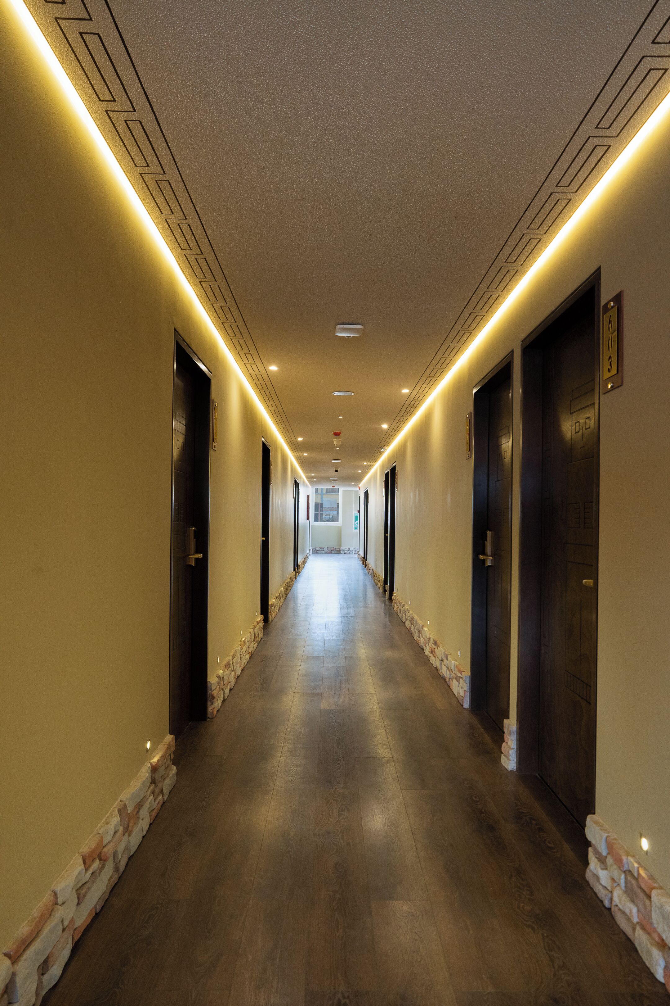 A long, narrow hallway with doors on both sides, leading to rooms. The hallway has a stone accent wall on one side and a plain wall on the other.