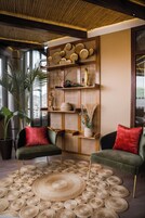 Una sala de estar moderna con un tapete tejido grande, dos sillones verdes con cojines rojos, una estantería de madera con objetos decorativos y una planta grande.