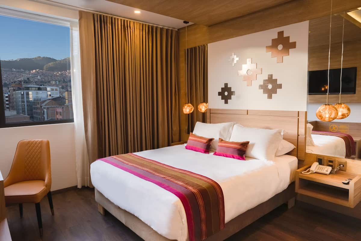 Quarto de hotel com uma cama grande, uma cadeira, uma mesa de cabeceira com telefone e vista da cidade pela janela.