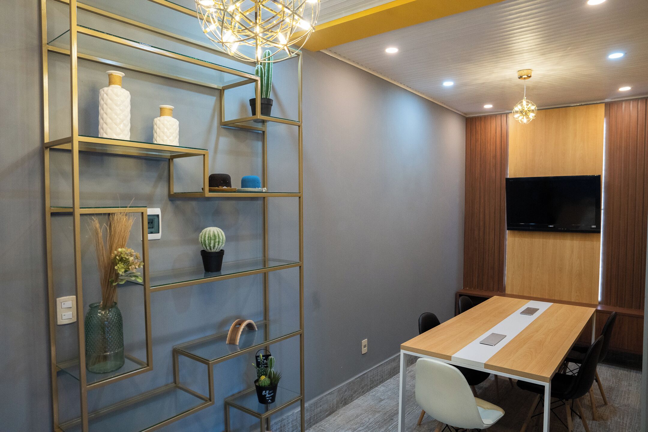 A modern meeting room with a wooden table, white chairs, a glass shelving unit with decorative items, and a flat-screen TV mounted on the wall.