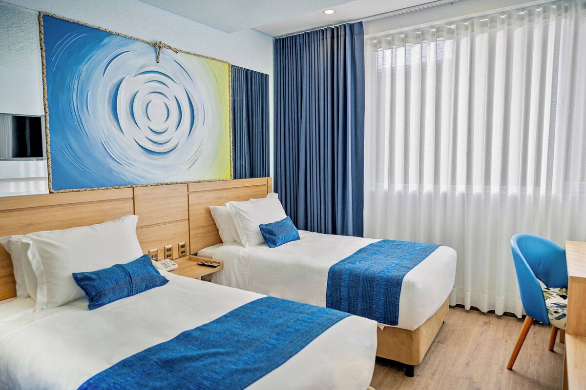 Quarto de hotel com duas camas, uma pintura abstrata grande, cabeceira de madeira, escrivaninha e uma cadeira.
