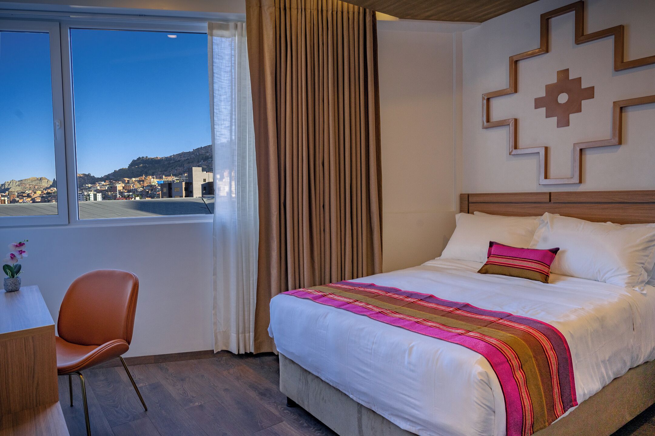A hotel room with a bed, a desk, and a chair. There is a window with a view of a cityscape.