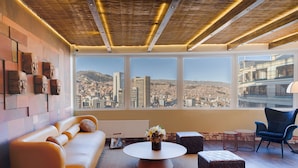 Ein modernes Wohnzimmer mit Sofa, Hocker, rundem Couchtisch und einem Sessel. Große Fenster bieten Blick auf die Stadt.