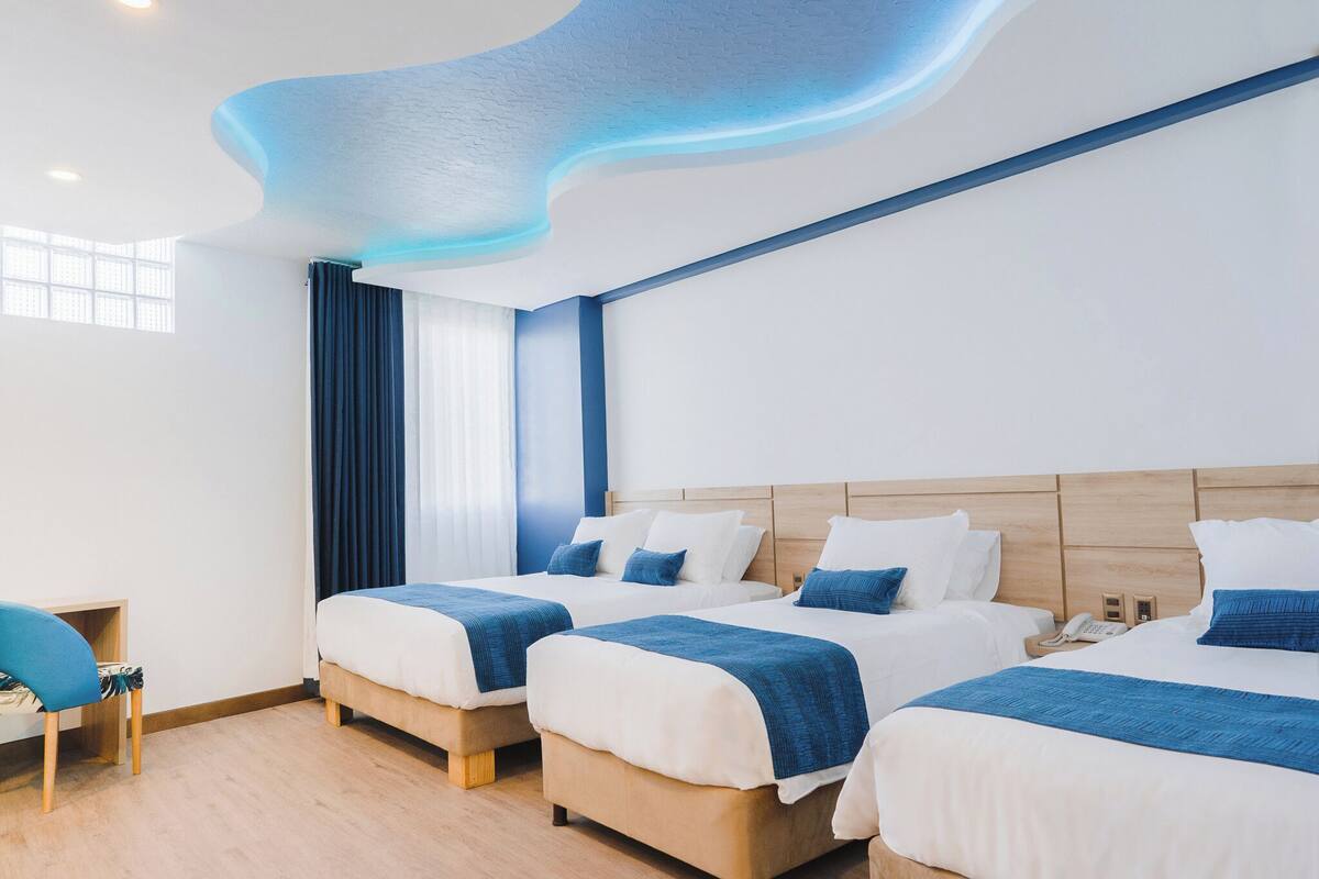 Quarto de hotel com três camas, uma escrivaninha e uma cadeira.