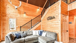 A cozy cabin interior with a wooden staircase, a sectional sofa, and a rustic decor including a deer skull and fish artwork.